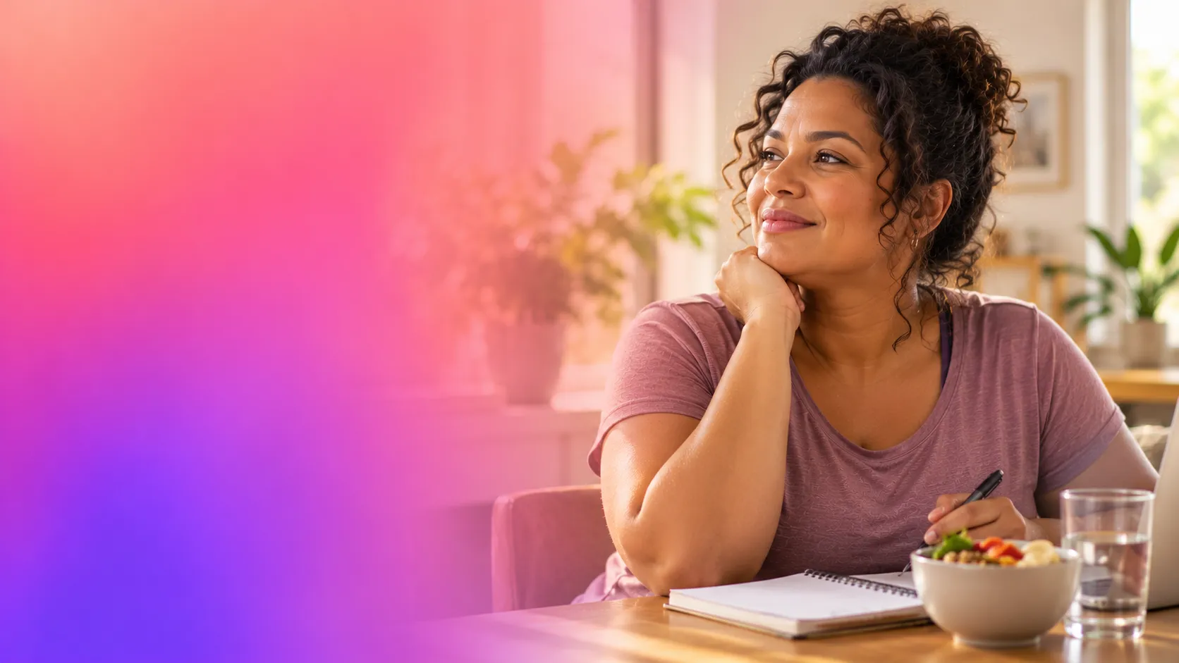 Mulher adulta de cabelos cacheados, sentada à mesa em um ambiente doméstico iluminado, com caderno, frutas e água à sua frente. À esquerda, degradê suave em tons de rosa, coral e roxo.